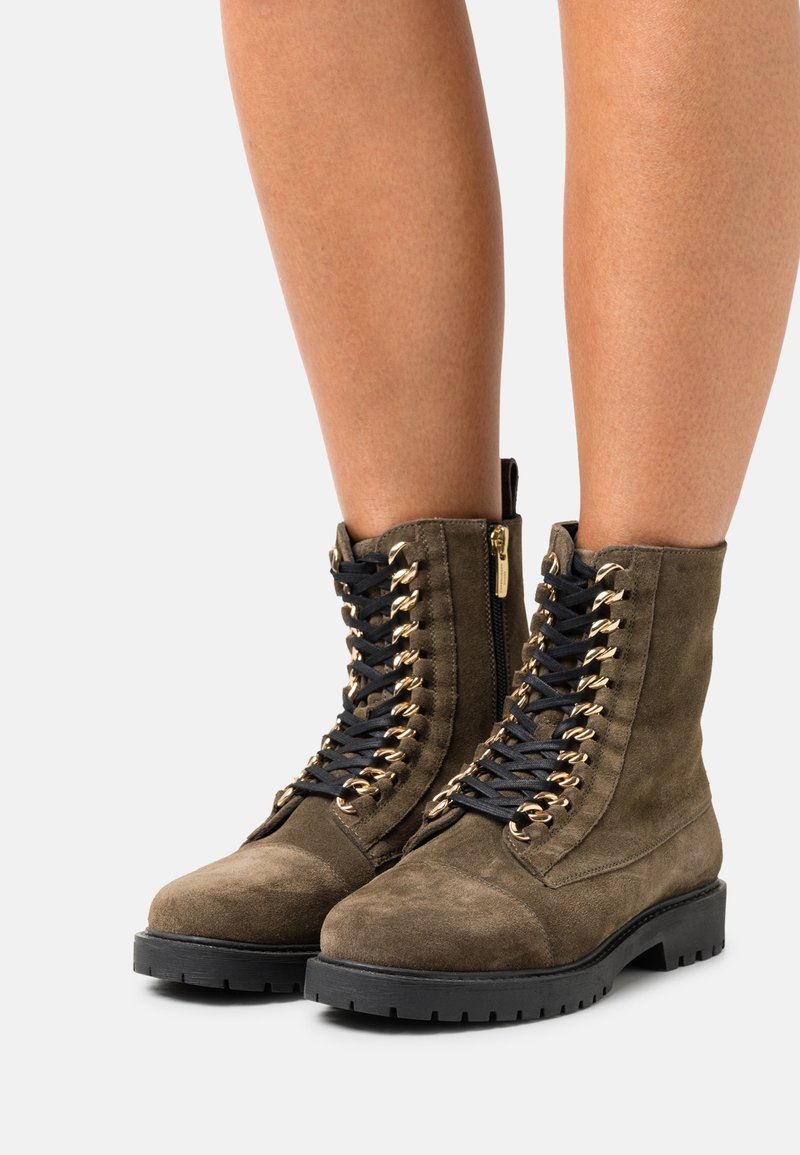 Bottes en daim olive montant à la cheville, avec des lacets noirs passant dans des œillets dorés et des semelles en caoutchouc noir, portées sur des jambes nues.