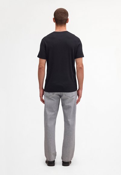 Mann steht mit dem Rücken zur Kamera, trägt ein schwarzes kurzärmeliges T-Shirt, hellgraue Jeans und schwarze Schuhe vor einem einfachen weißen Hintergrund.