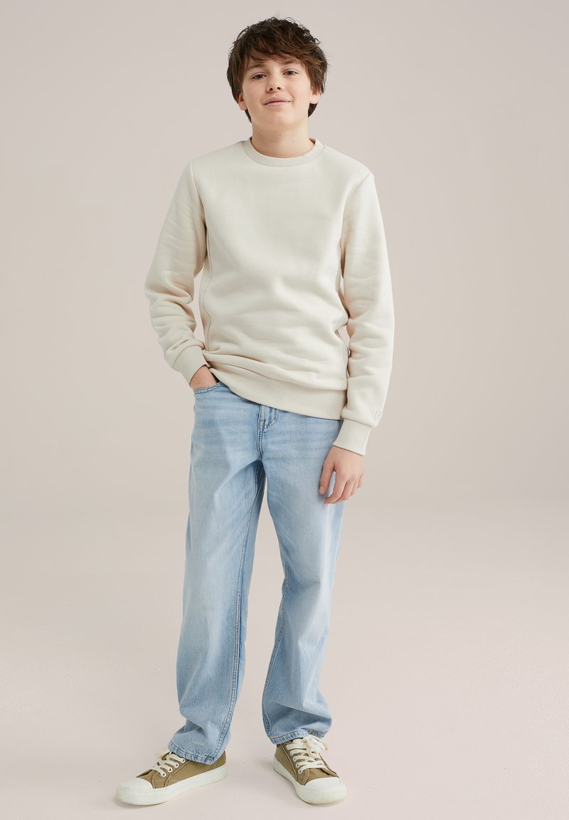 Svetlo bež sweatshirt s rebrastimi manžetami, v kombinaciji s svetlo modrimi hlačami ravnega kroja. Obutev ima bele podplate in zgornji del iz olivno zelenega platna.