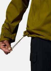 Hand adjusts drawstring on olive green jacket cuff, showing sleeve and black cargo pants pocket against light background.