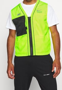 Nike-reflekterande väst i klar neongrön, med en svart nätficka, framdragkedja och logodetalj. Lättviktigt tyg med slät textur.