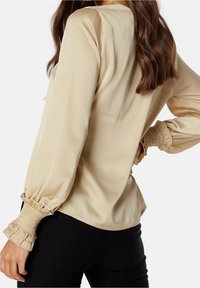 Blouse en satin beige avec manches bouffantes, poignets froncés et encolure ronde. Texture lisse, polyvalente pour diverses occasions.