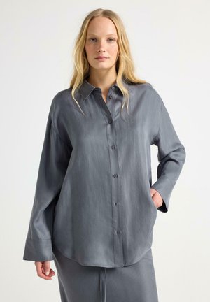 Kvinde med langt blondt hår iført løs grå skjorte med knapper og matchende nederdel, stående mod en ensfarvet lys baggrund.