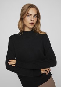 Fekete kötött pulóver magas nyakkal, bordás mandzsettákkal és szegéllyel, sima textúrával, testhez simuló sziluettel és hosszú ujjakkal.