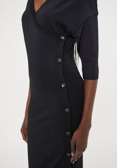 Schwarzes Strickkleid mit V-Ausschnitt, kurzen Ärmeln und seitlichen Knopfdetails. Die glatte Textur und die figurbetonte Silhouette betonen das Design.