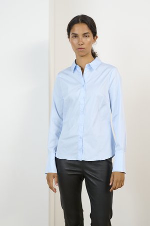 Camicia blu chiaro con colletto e maniche lunghe; presenta una texture liscia e una vestibilità dritta, abbinata a pantaloni in pelle nera.