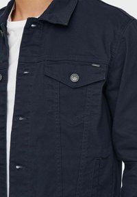 Giacca in denim navy con chiusura a bottoni, due tasche sul petto e una finitura testurizzata. Include accessori in argento e un design classico del colletto.