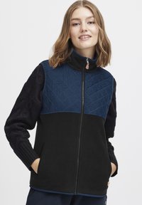Gilet en polaire avec un corps noir, un empiècement matelassé bleu marine et des manches texturées foncées avec un motif en maille torsadée. Fermeture éclair à l'avant et poches latérales.