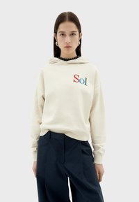 Beige hooded sweatshirt with "Sol" embroidered in red and green. Features ribbed cuffs and hem, paired with navy wide-leg trousers.