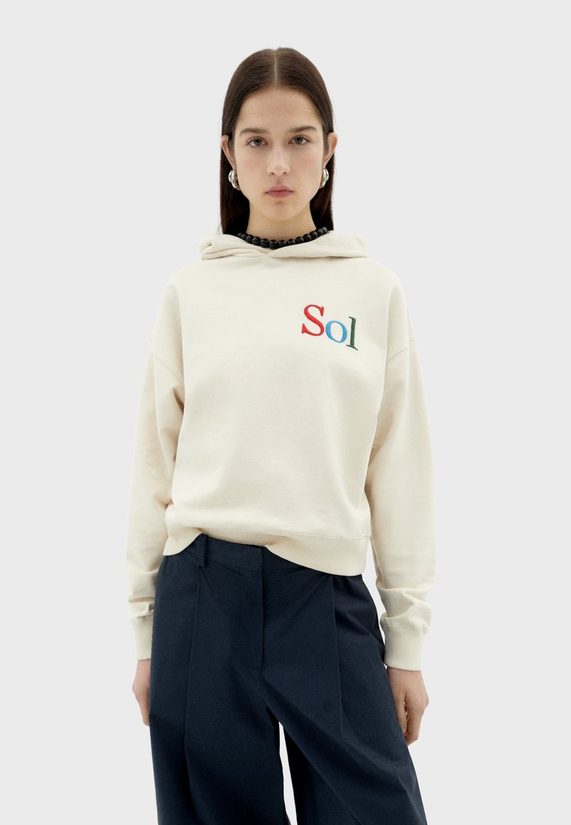 Beige hooded sweatshirt with "Sol" embroidered in red and green. Features ribbed cuffs and hem, paired with navy wide-leg trousers.