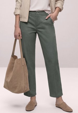 Persona con pantalones verdes de pierna recta, chaqueta beige y pantuflas, sosteniendo un gran bolso tote de ante marrón claro con una mano en el bolsillo.