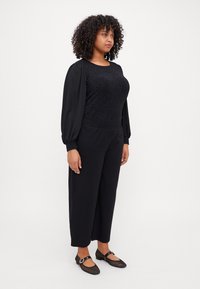 Black textured top with puff sleeves, paired with high-waisted black trousers and mesh detail shoes with a strap and buckle.