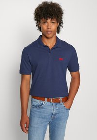 Polo shirt blu navy in tessuto di cotone con colletto classico, maniche corte e un piccolo logo rosso sul petto, abbinato a jeans azzurri.