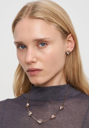 Goldfarbene Schmuckstücke mit herzförmigen Anhängern aus transparentem Material. Das Model trägt passende Ohrringe und ein enges dunkles Oberteil.