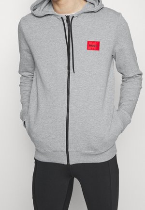 Felpa grigia con zip realizzata in tessuto morbido. Presenta un piccolo logo quadrato rosso sul petto, cappuccio con cordino e tasche frontali. Polsini a coste testurizzati.