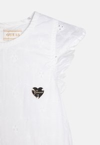 Vit ärmlös topp med volangdetaljer vid axlarna, blommig broderad dekoration och en svart hjärtformad Guess-logotyp som accent.