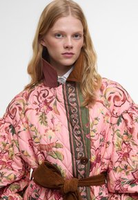Prošívaný růžový kabát s květinovými vzory, zelenými akcenty a hnědým kordovým límcem. Obsahuje pásek a dekorativní švy.