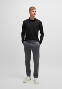 Čierna polo košeľa s dlhým rukávom a golierom, kombinovaná so sivými nohavicami a béžovými teniskami, vytvára jednoduchý, moderný outfit.
