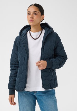 Femme portant une veste matelassée à capuche bleu foncé sur une chemise blanche et un jean bleu, avec un collier de perles marron et des boucles d'oreilles dorées.