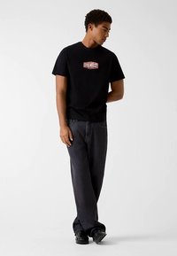 Zwart T-shirt met korte mouwen en een rechthoekig rood-wit logo op de borst. Gecombineerd met donkere spijkerbroek en zwarte schoenen.