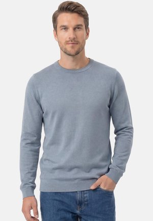 Homme aux cheveux courts portant un pull bleu clair à col rond et un jean bleu, debout avec une main dans la poche devant un fond uni.