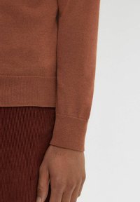 Maglione marrone a maglia con dettagli a coste sui polsini, realizzato in materiale morbido. La mano si appoggia su un tessuto di velluto a coste borgogna testurizzato.