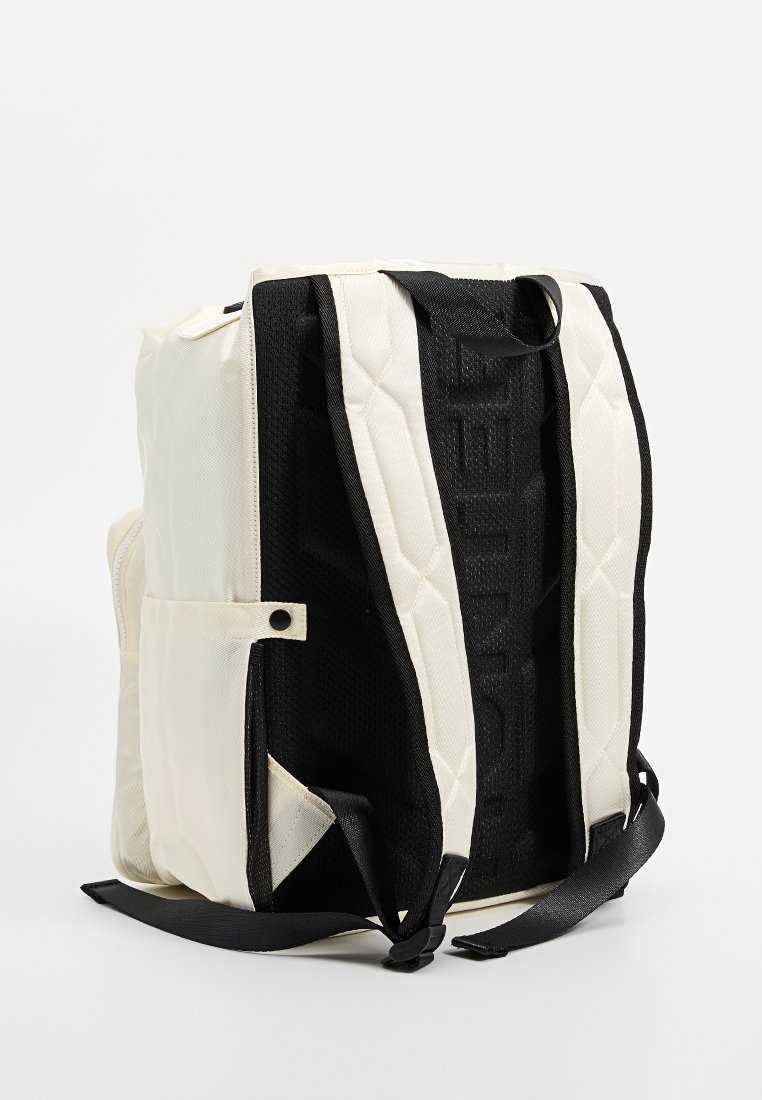 Mochila blanca y negra con textura acolchada, cuenta con correas ajustables, bolsillos laterales y un respaldo de malla. Diseño minimalista.
