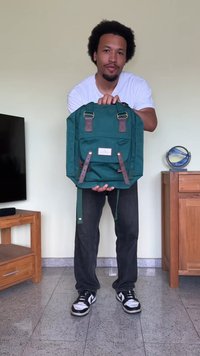 Mochila azul-petróleo com detalhes em couro castanho, ferragens metálicas, bolso frontal e alças ajustáveis, segurada por uma pessoa vestida de forma casual.