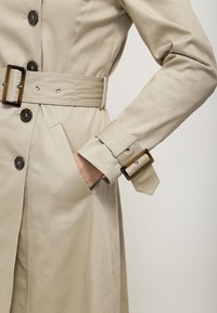 Gros plan sur un trench beige avec des boutons foncés, une ceinture avec une boucle en écaille, une sangle au poignet et une main dans la poche latérale.