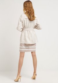 Beżowy trencz z paskiem w talii, z długimi rękawami i bocznymi kieszeniami, w połączeniu z beżową spódnicą w białe poziome paski.