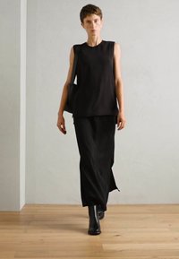 Top sin mangas negro combinado con una falda larga y fluida negra. La modelo lleva botines negros hasta el tobillo y carga un bolso negro de hombro. Textura suave.