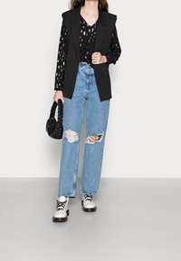 Schwarzer Nadelstreifen-Blazer über einem gemusterten schwarzen Oberteil, kombiniert mit hellblauen, verwaschenen weit geschnittenen Jeans und weißen Schnürschuhen. Hält eine schwarze geflochtene Tasche.