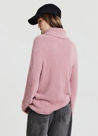 Maglione rosa peloso con collo alto, maniche lunghe e vestibilità rilassata. Indossato con pantaloni grigio scuro e un berretto nero.