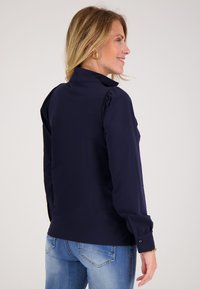 Vrouw met blond haar, gekleed in een marineblauwe blouse met lange mouwen en lichtblauwe jeans, glimlachend en naar rechts kijkend tegen een effen achtergrond.