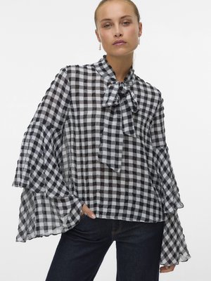Žena v černo-bílé kostkované blůze s mašlí u krku a rozšířenými rukávy, kombinuje ji s tmavě modrými džínami.