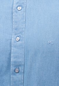 Ανοιχτό μπλε πουκάμισο με λευκά κουμπιά και μικρό λευκό κέντημα με το λογότυπο "MK" στην περιοχή του στήθους.