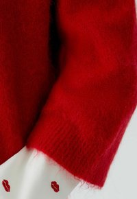 Roter Strickpullover mit flauschiger Textur; ausgestattet mit gerippten Bündchen und kontrastierendem weißen Stoff darunter, verziert mit roter Lippenstickerei.