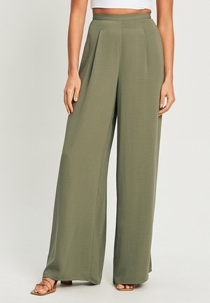Vrouw draagt een olijfgroene broek met hoge taille en wijde pijpen, gecombineerd met beige open-teen sandalen met hakken en een wit cropped topje.