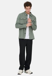 Mann, der den Ärmel eines olivgrünen Hemds mit Knöpfen anpasst, trägt weißes T-Shirt, schwarze Hose und beigefarbene Turnschuhe, steht vor weißem Hintergrund.