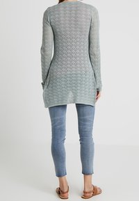 Tunique en tricot bleu clair à manches longues avec un motif en diamant texturé, associée à un jean slim-fit bleu clair et des sandales marron.