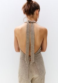 Beige, textured jumpsuit with an open back and tie detail at the neck and waist. Smooth fabric showcases a subtle pattern.