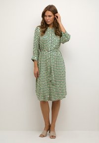 Rochie verde cu un model geometric de triunghiuri în albastru și mov. Are mâneci lungi, cordon și o lungime până la genunchi. Este completată de pantofi bej.