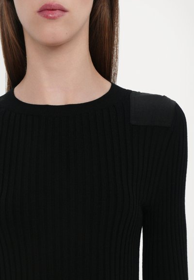 Black ribbed knit top featuring a round neckline and a smooth fabric patch on the left shoulder. The overall texture is soft and fitted.