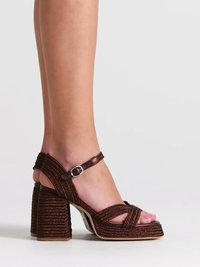 Jambes portant des sandales à talons hauts tressées marron avec des brides à la cheville et des boucles argentées sur fond blanc.