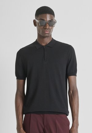 Camiseta polo negra de manga corta con cremallera frontal, hecha de un tejido suave. Cuenta con un cuello clásico y puños de manga acanalados. Se lleva con pantalones de color burdeos oscuro.