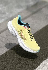 Baskets de course jaunes vives avec une tige texturée, des lacets noirs et le logo HOKA. Présente des accents bleu clair et une semelle intermédiaire blanche épaisse.