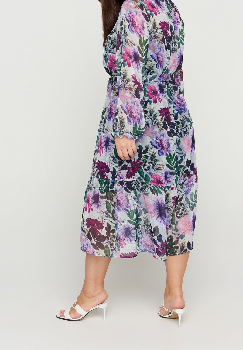 Femme portant une longue robe à fleurs violettes, roses et vertes, associée à des sandales blanches à talons hauts, debout de profil.