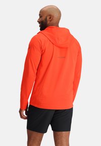 Rode lichtgewicht jas met kap, gestructureerd oppervlak en logo op de achterkant. Draagt bij zwarte shorts.