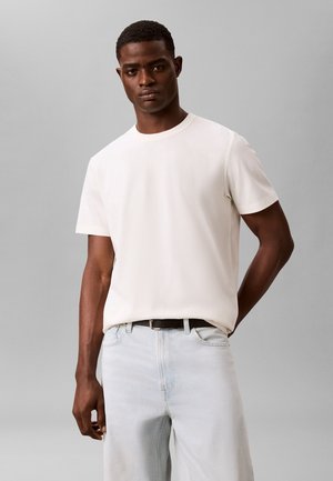 Homme portant un t-shirt blanc uni à manches courtes et un jean bleu clair, debout devant un fond gris uni.