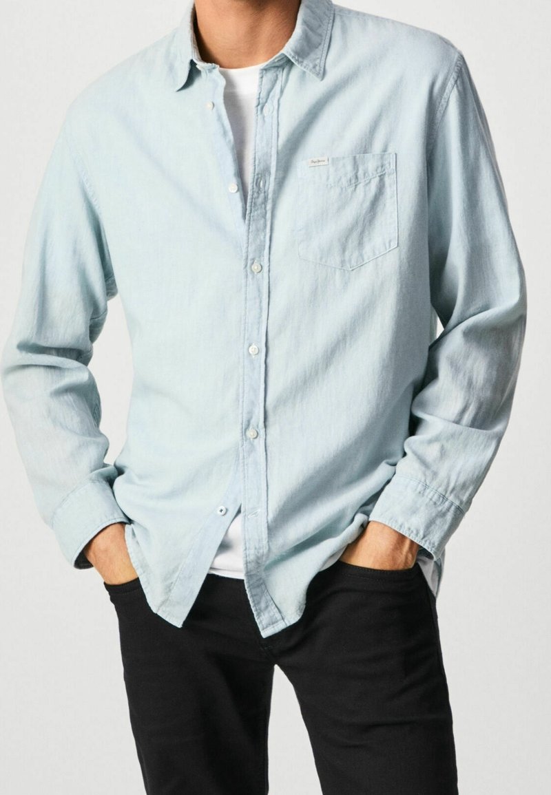 Camicia button-up a maniche lunghe azzurro chiaro in un tessuto morbido, con una tasca sul petto e una vestibilità rilassata. Indossata sopra una camicia bianca.